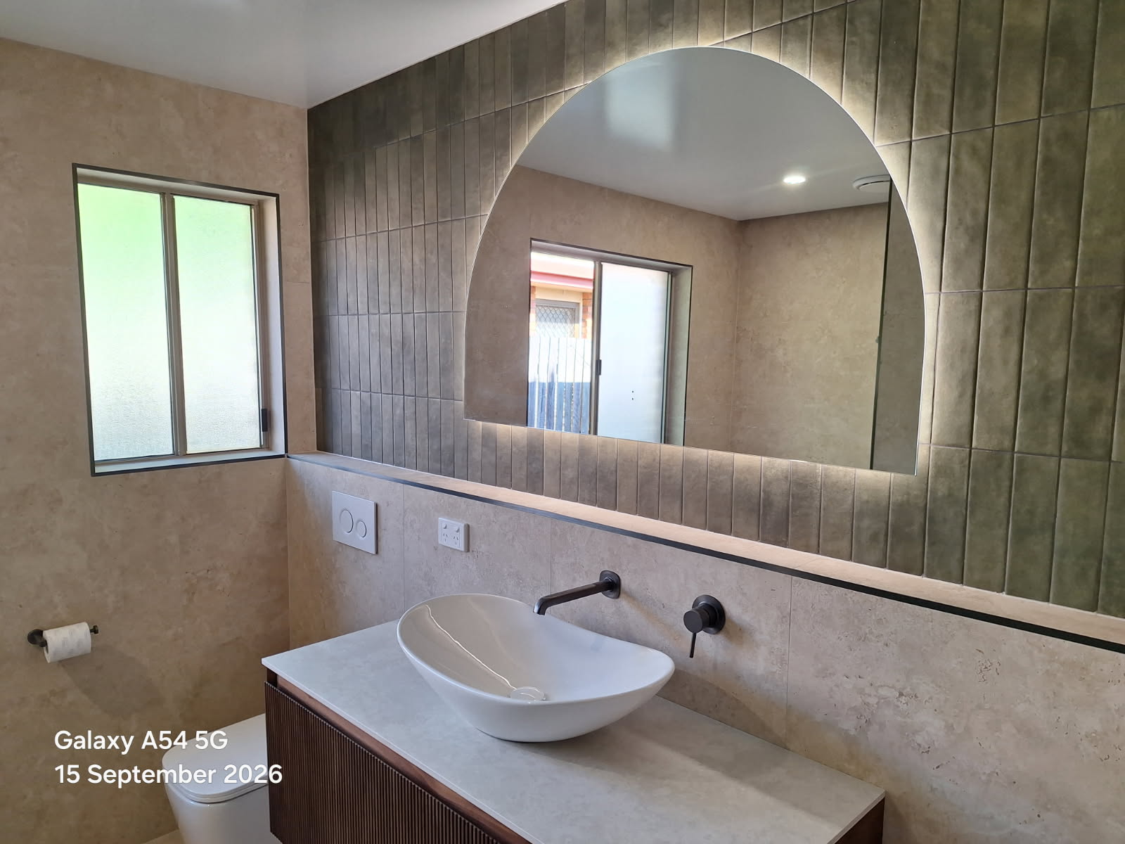 5 Modern bathroom with a circular white vessel sink on a stone countertop, a large arched backlit mirror, and beige tile walls.