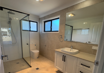 Modern bathroom with a glass-enclosed shower, toilet, white vanity with an oval sink, large mirror, beige tiling, and two rectangular windows.